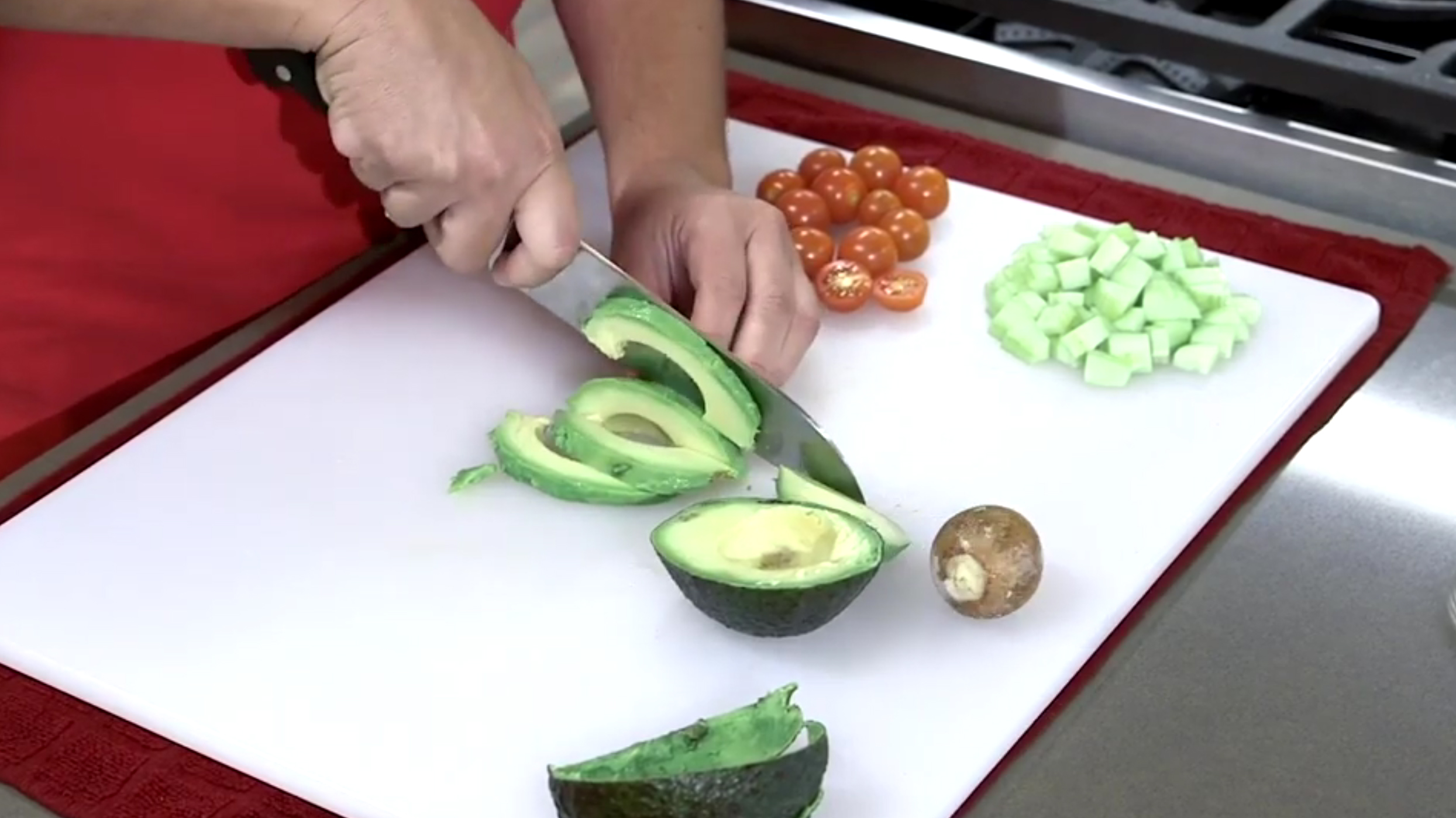 Hand with a knife slicing an avocado on a cutting board alongside diced cucumbers and cherry tomatoes.
