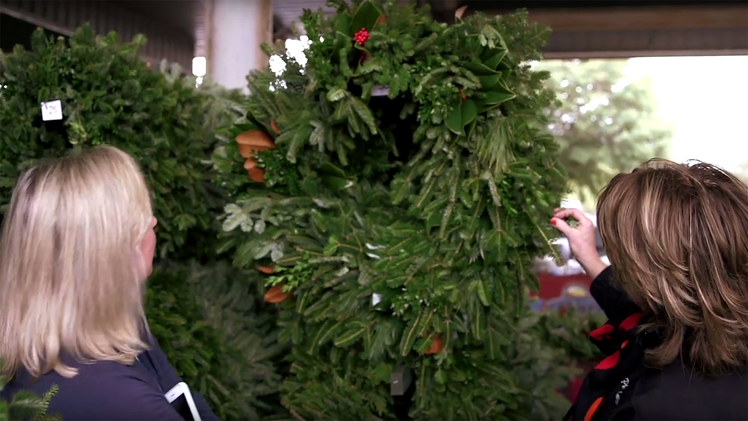 Two women looking at a green holiday wreath hanging on a rack.