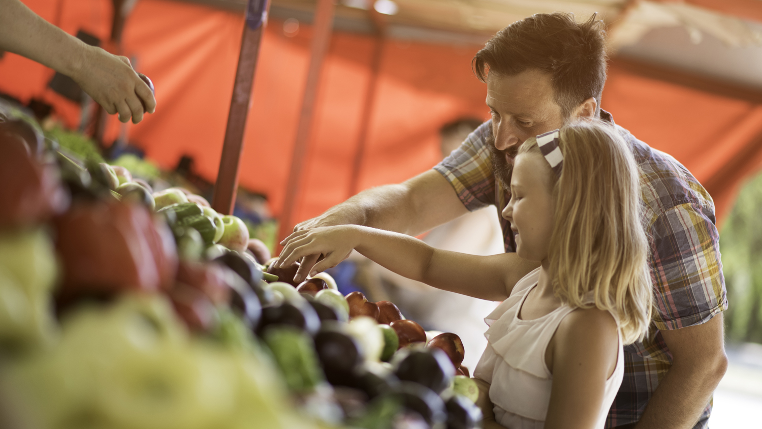 Smiling bearded man and his daughter buying fresh produce at outdoor farmers market stand.