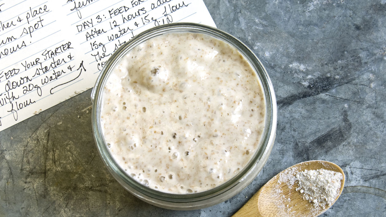 Flat lay photo of a jar of sourdough starter on a counter with a recipe and a spoon.