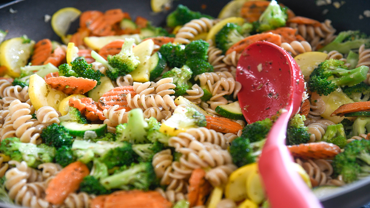 A skillet with whole wheat pasta, vegetables, and a spoon.