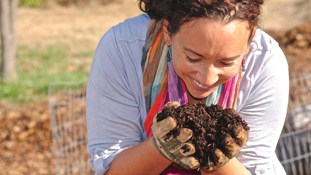 A smiling woman examines soil up close in her hands while gardening.