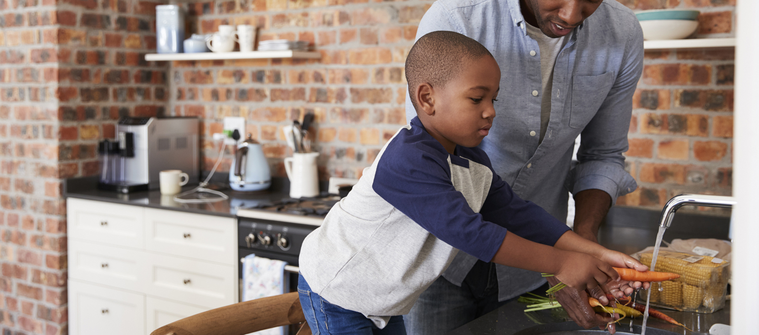 Young boy helping his father wash carrots under running water in kitchen sink