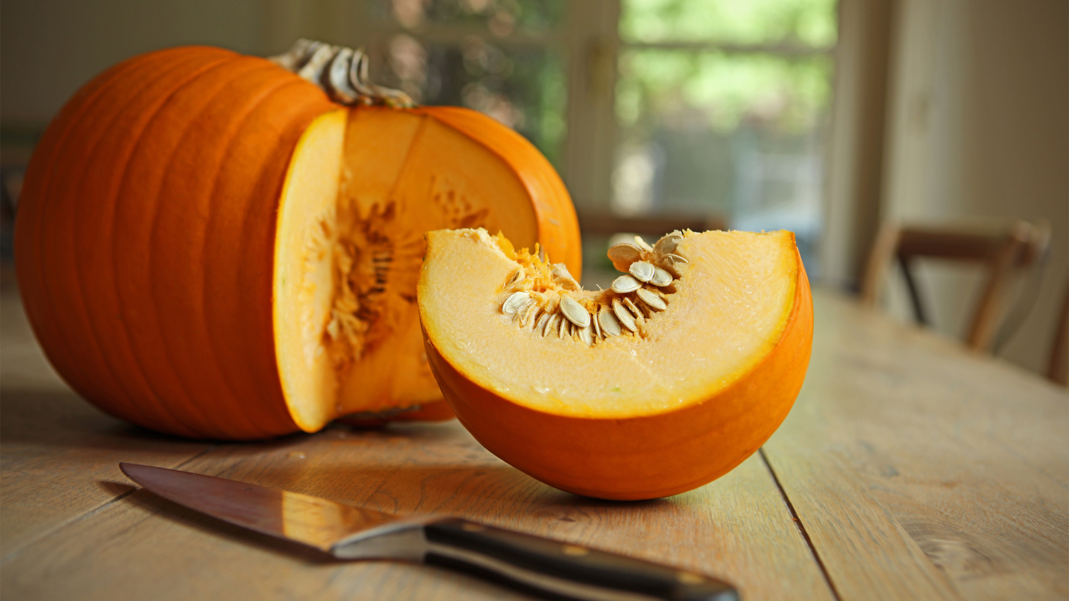 An orange pumpkin on a kitchen table with a slice cut off and laying next to the pumpkin, as is a kitchen knife.