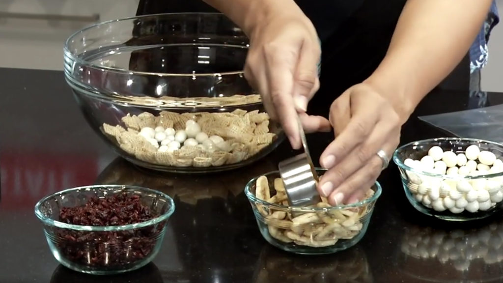 Close up of hands scooping out dried bananas, alongside glass bowls of other healthy snacks like raisins, to make a toss up snack bag.