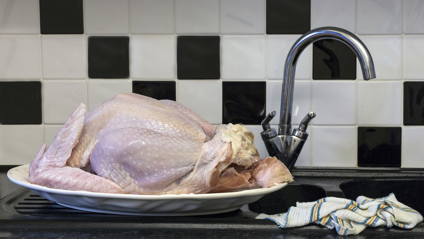 Raw whole turkey defrosting on a kitchen sink. A dish cloth is in close proximity to the defrosting meat, creating a cross contamination and food safety issue.