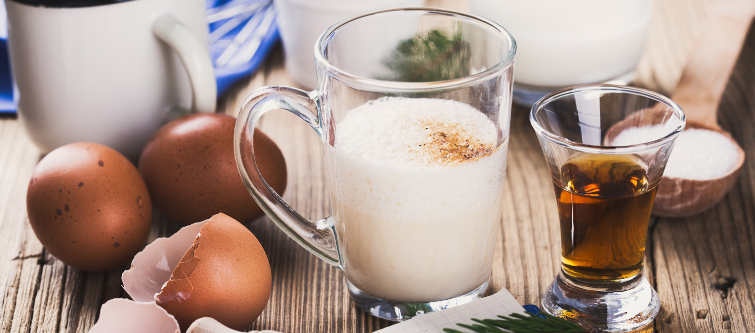 A cup with eggnog surrounded by ingredients like eggs and a recipe book on a rustic wooden table