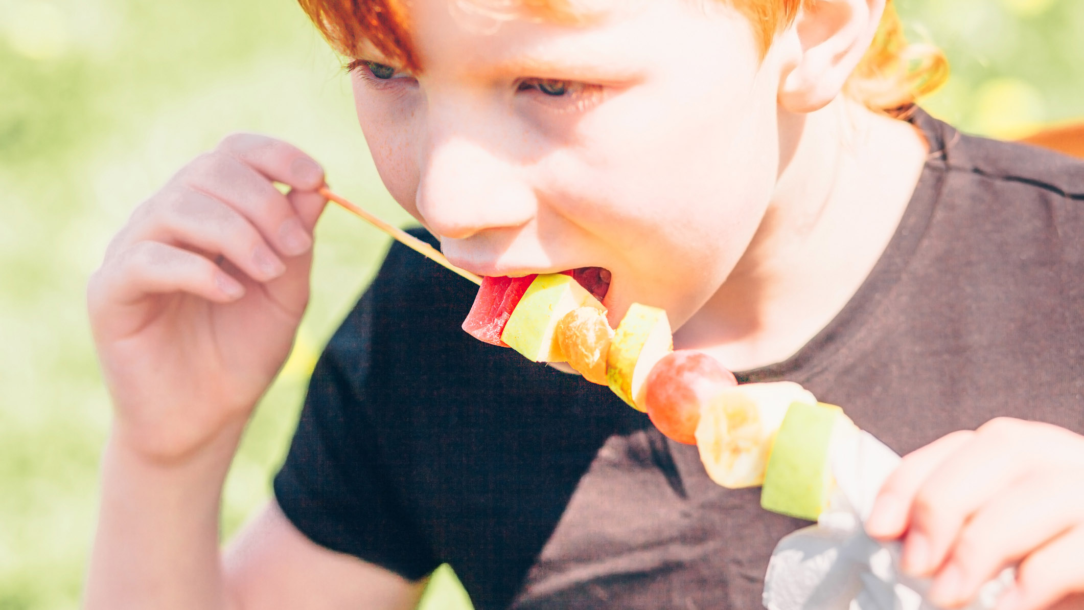 Red-haired boy eating a fruit kabob with berries and other fresh fruit on a wooden skewer
