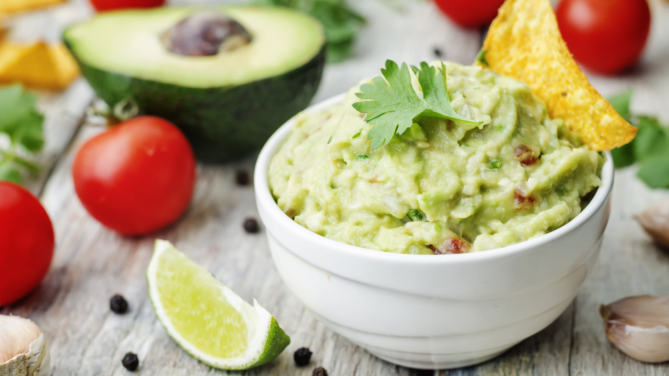 Guacamole with corn chips on a white wood background, surrounded by small tomatoes and a lime wedge