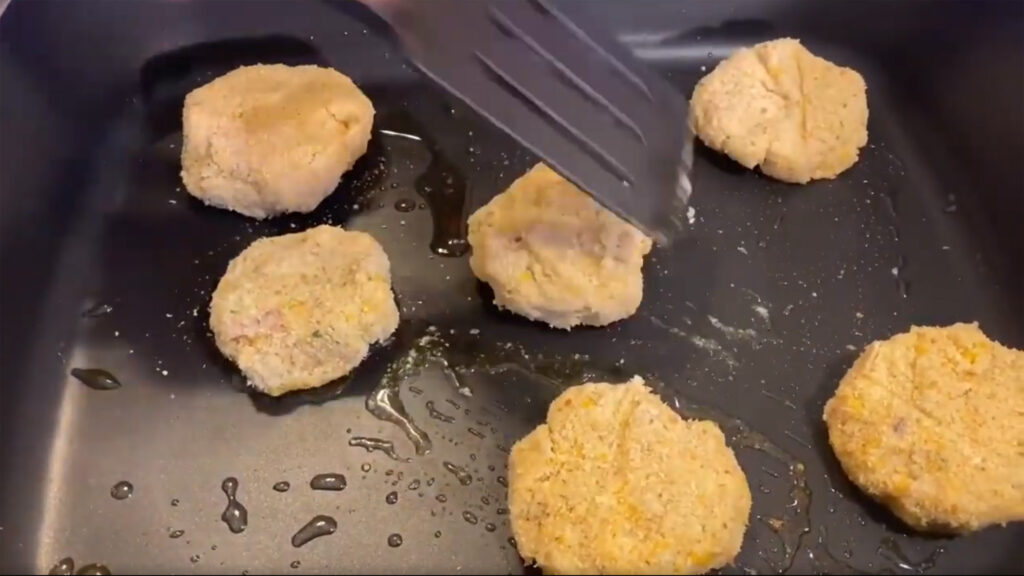 Tuna patties cooking on a skillet, while a spatula is preparing to flip one of them.
