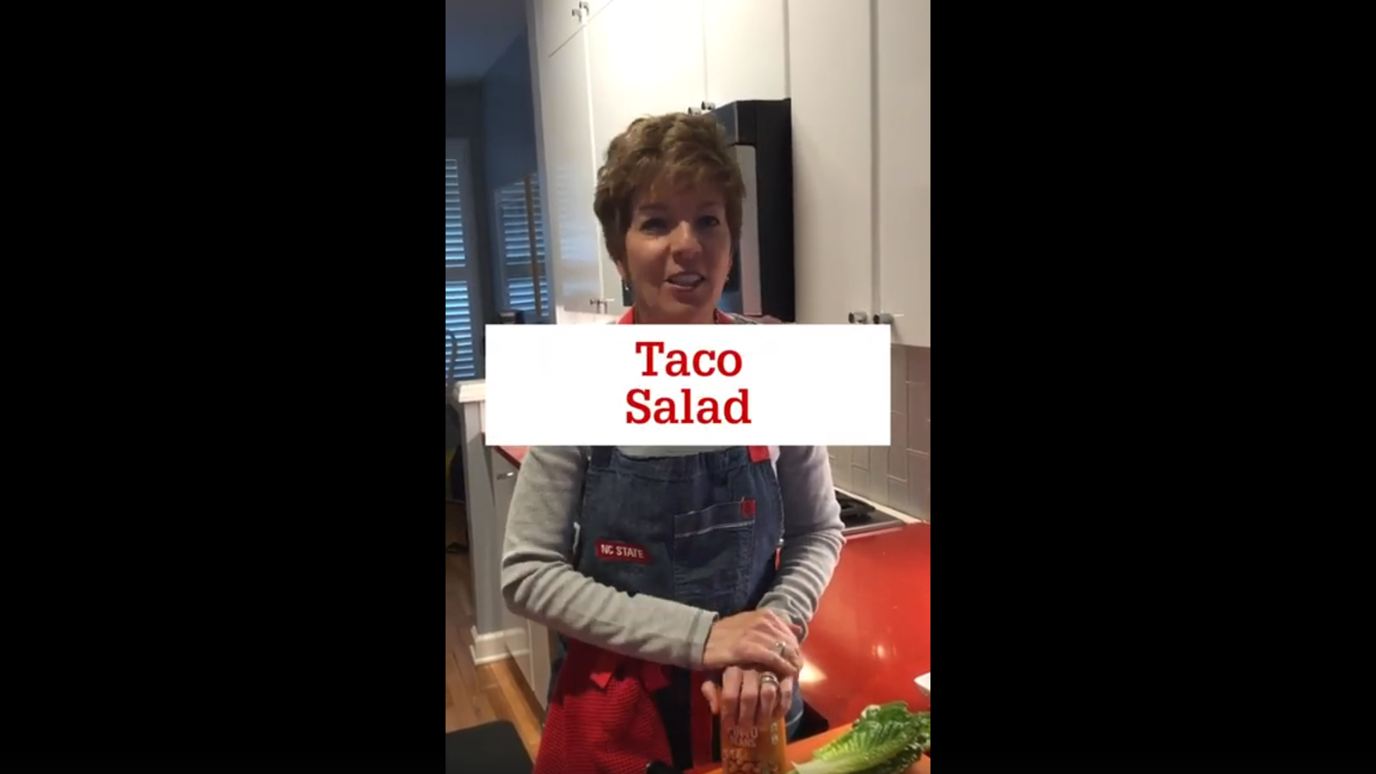 A woman wearing an apron is smiling while standing at her kitchen counter with ingredients for a taco salad laid out. The words "Taco Salad" are displayed across the image.