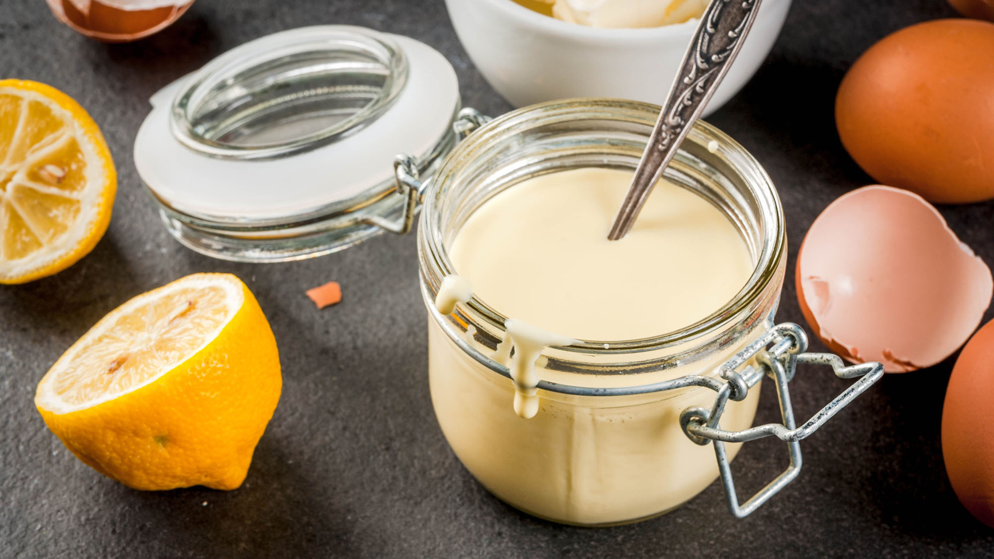 Hollandaise sauce in a glass serving jar, with ingredients like eggs and lemons around the jar on a black stone table.