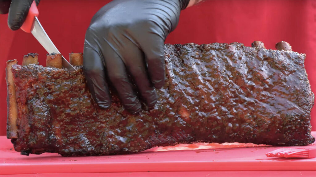 Close shot of gloved hands holding and cutting through barbecue spare ribs on a red tabletop.