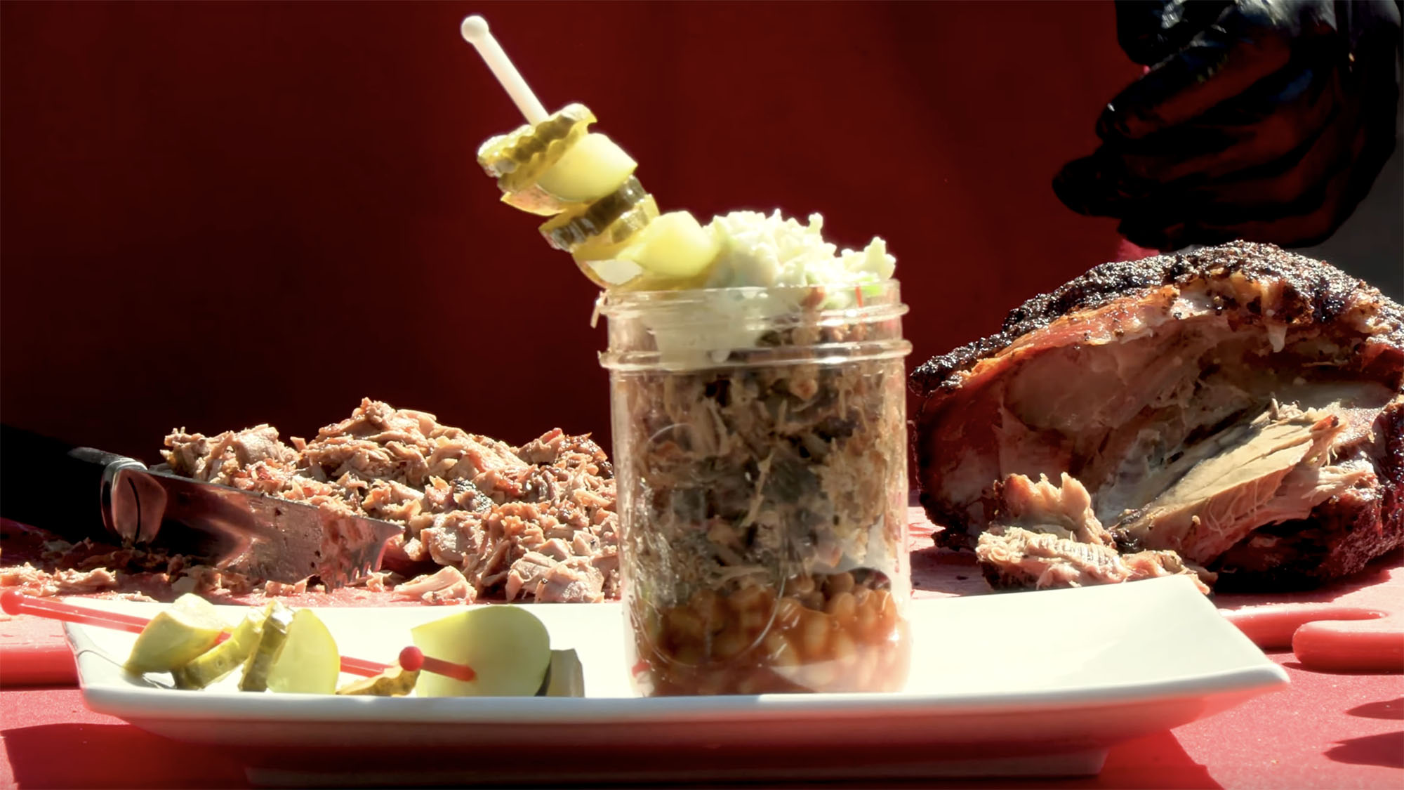 A glass jar with a "barbecue martini" consisting of pork barbecue, baked beans, cole slaw and pickles sits on a white plate while displayed on a red tabletop outdoors.