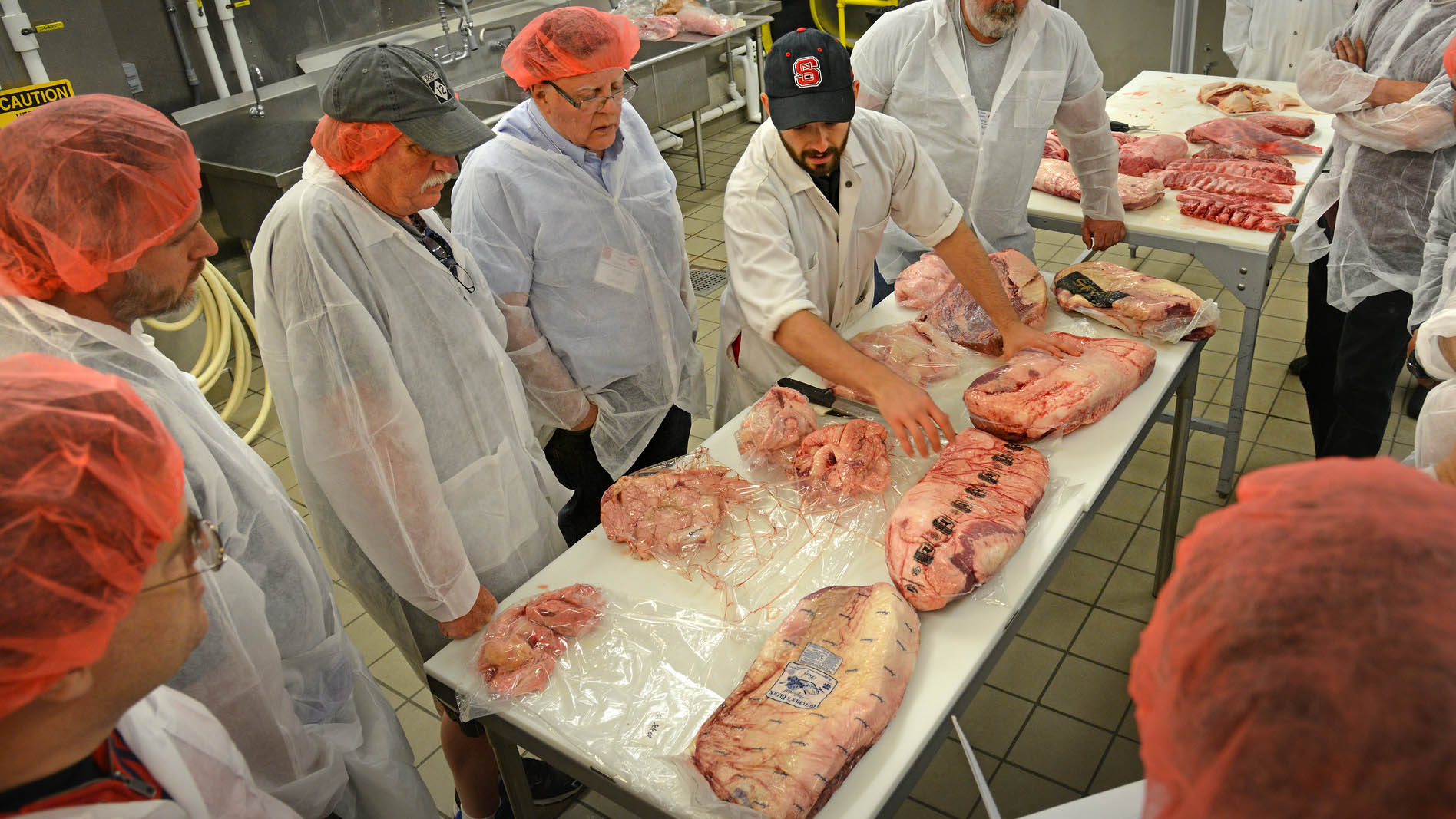 Participants wearing hair nets and food safety coverings gather around a table of fresh cut meats as part of NC State University's BBQ Camp.