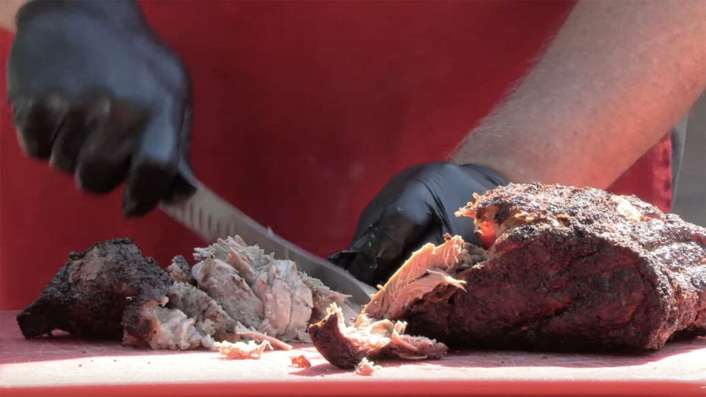 Close shot of gloved hands holding and cutting through cooked pork butt barbecue on a red tabletop.