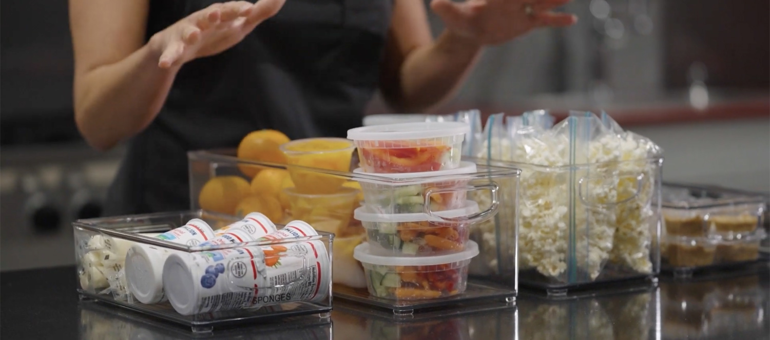 Plastic storage bins are displayed side by side on a kitchen countertop, each of which is filled with various snack items like cheesesticks, yogurt, fruit cups, nuts and other items.