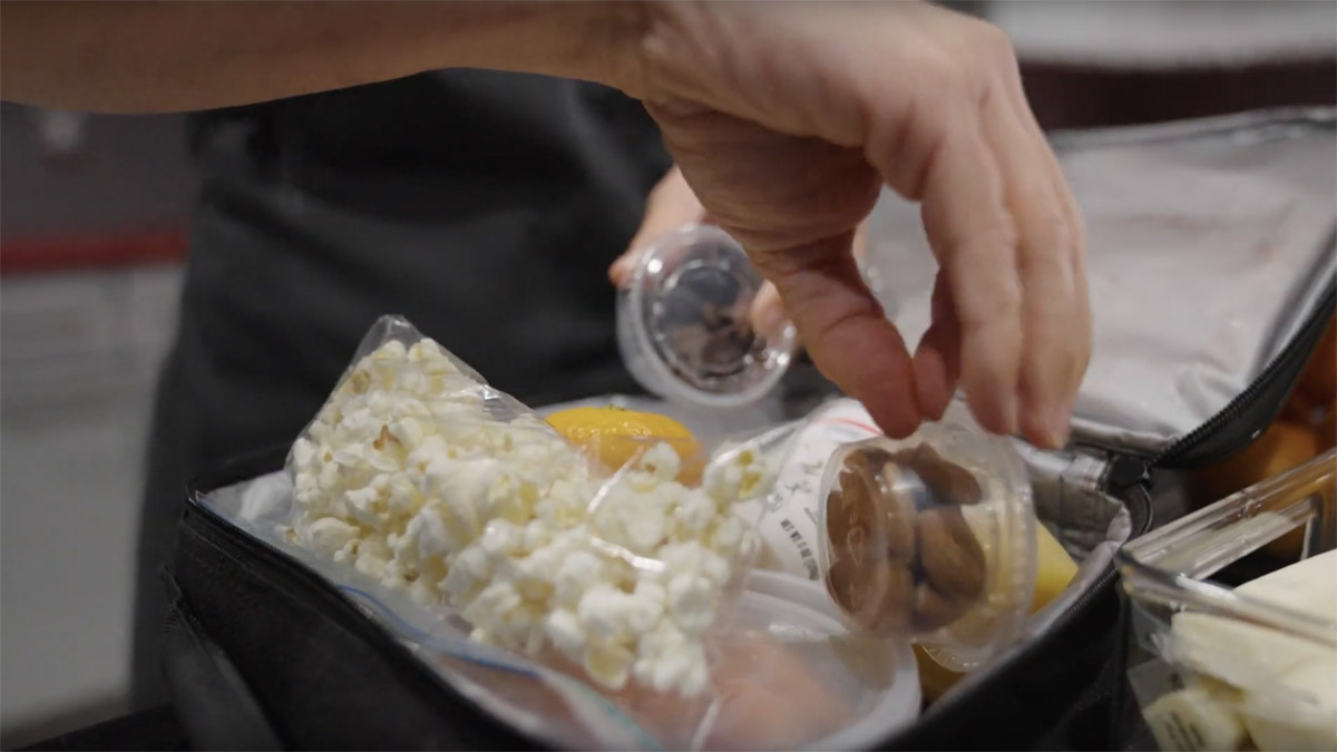 A hand is shown placing healthy snack items, like bagged popcorn, a container of nuts and yogurt, into a child's lunchbag for school.