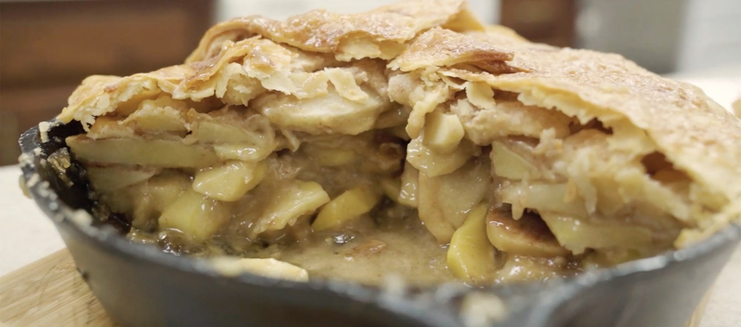 A fully-baked mountain high apple pie still in the skillet with a slice cut out showing the apples and ooey gooey goodness inside.