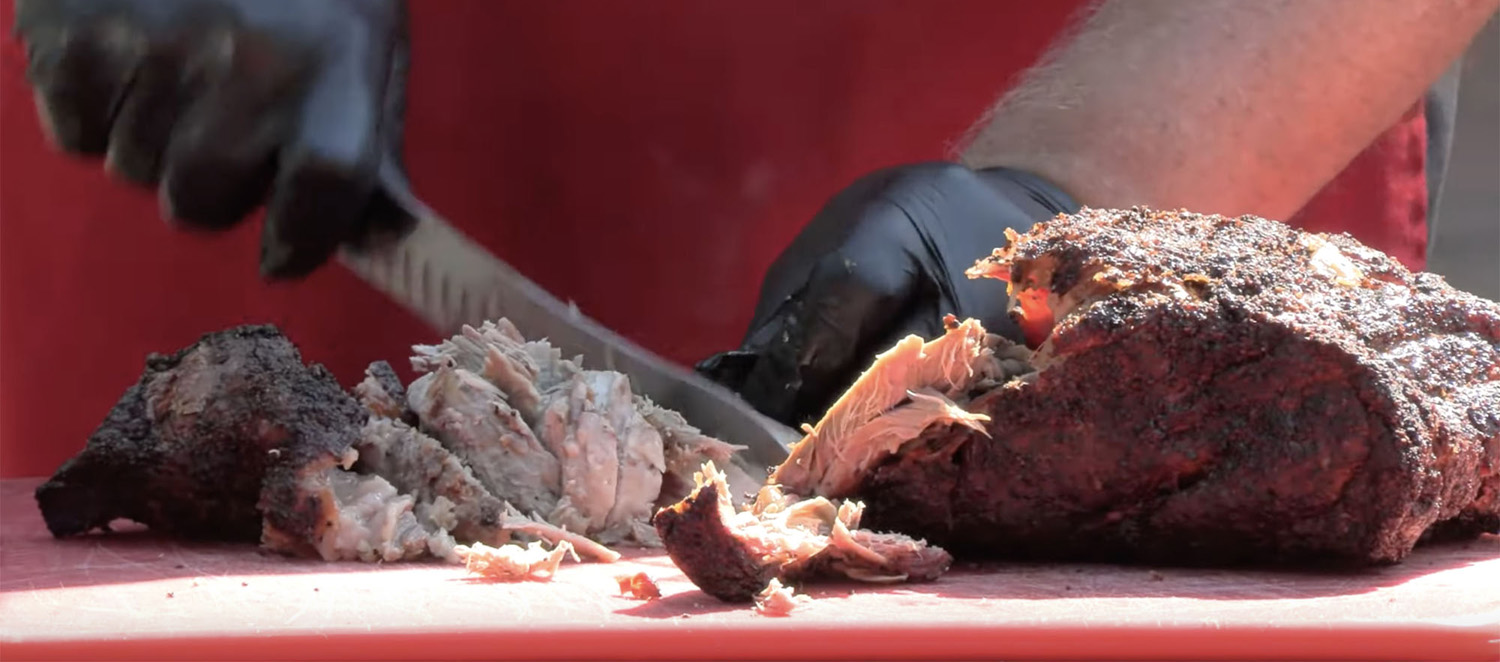 A pair of gloved hands are holding and cutting through cooked pork butt barbecue on a red tabletop.