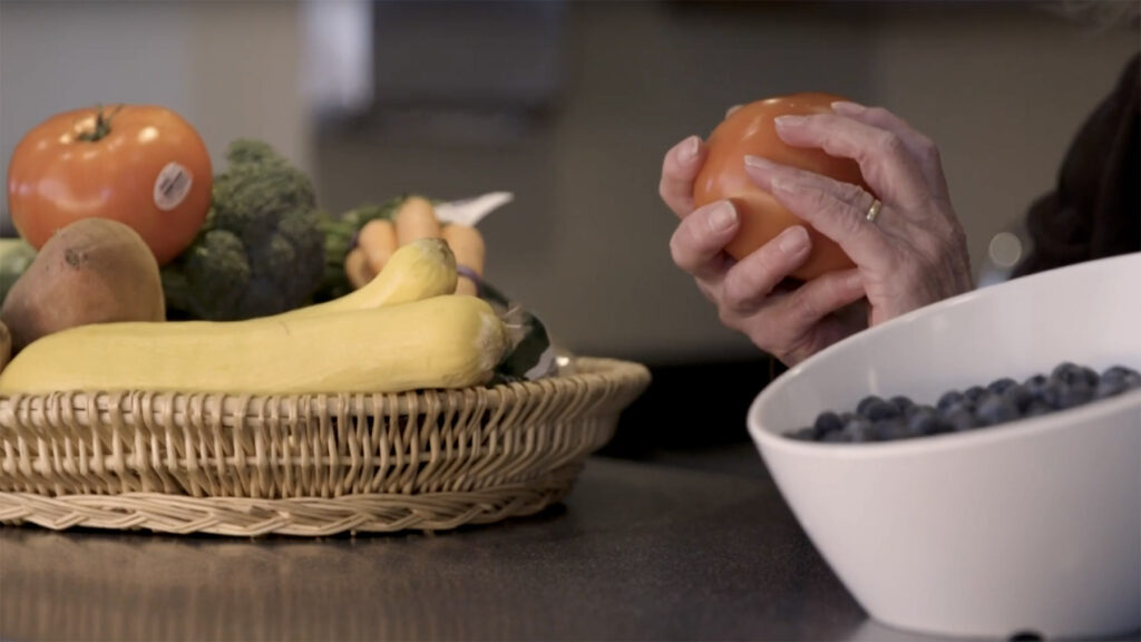Countertop with bowls of fresh fruits and vegetables, like bananas, blueberries and broccoli, with hands holding a tomato in the background.